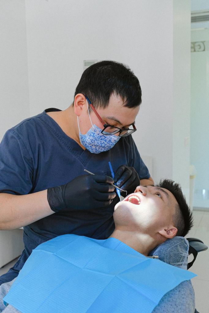Dentist in scrubs and mask examines patient’s teeth while the patient lies back with mouth open in a dental chair.