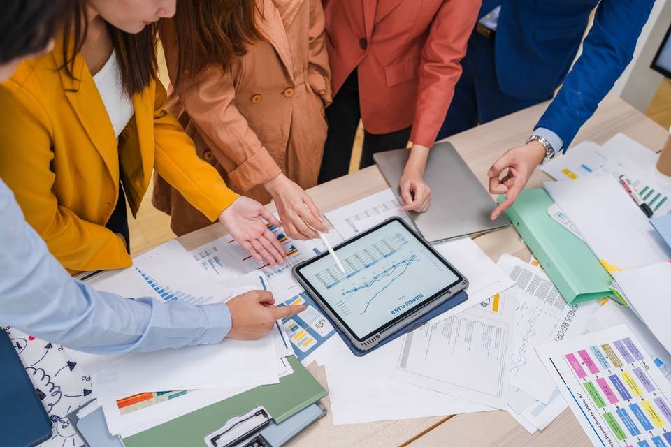A close-up of several people standing around a desk covered with papers. They're all pointing at a tablet displaying a graph. - Lead Grow Develop - Business, Technology, Personal Development A close-up of several people standing around a desk covered with papers. They're all pointing at a tablet displaying a graph.