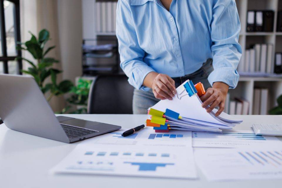 Person in a blue shirt stacks and clips papers with colorful binder clips at a white desk, laptop nearby.