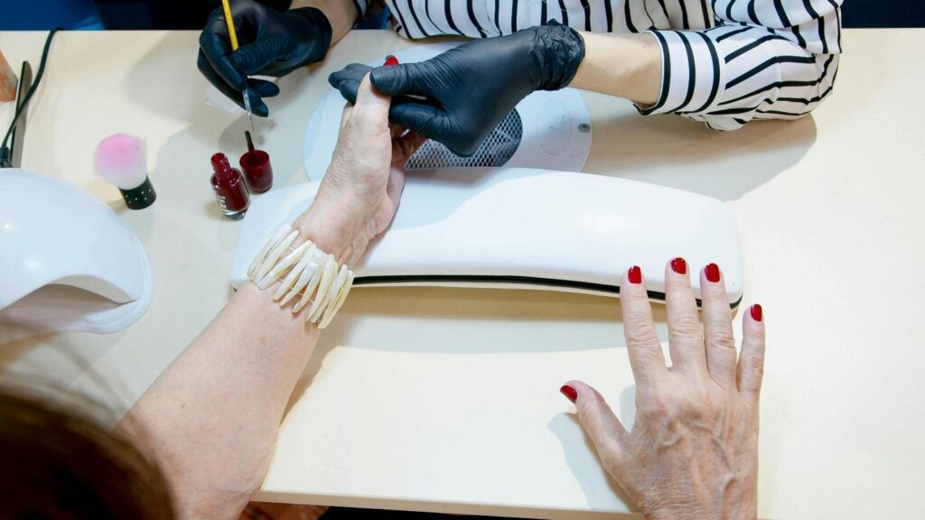 Manicurist in black gloves paints red nail polish on a client's fingernails at a nail salon station.