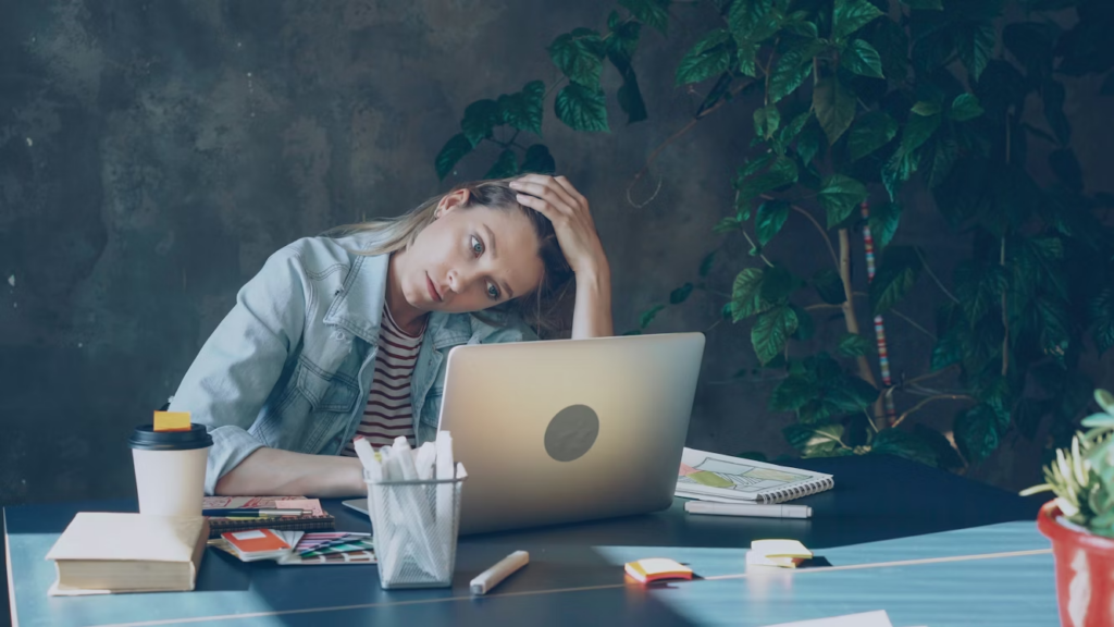 Woman sits at a desk with a laptop, head resting on her hand, looking stressed; coffee cup and notebooks on the table with green plants in the background.