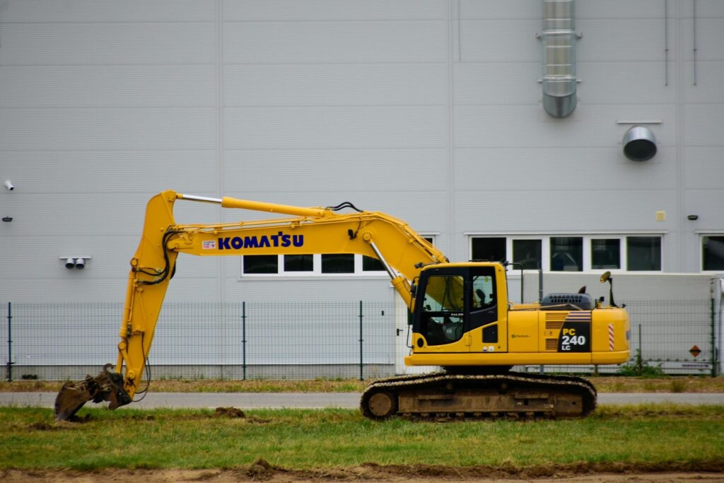 Yellow Komatsu excavator at a construction site in front of a gray industrial building with a chain-link fence behind it.