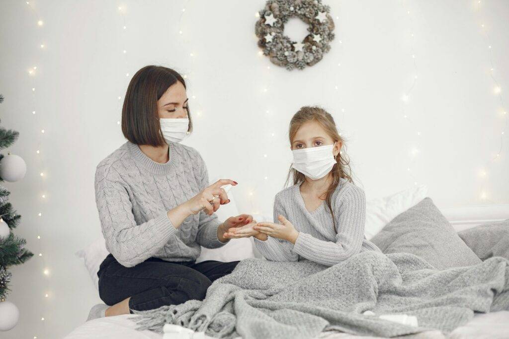Mother and daughter wearing masks, seated together on a bed with a gray blanket, crafting with small objects as warm white string lights and a wreath glow in the background.