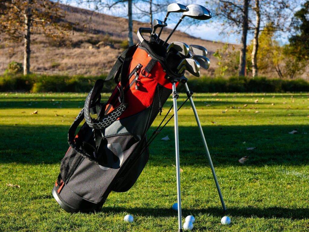Golf bag with several irons standing on a golf course, white golf balls scattered on the grass in front of it.