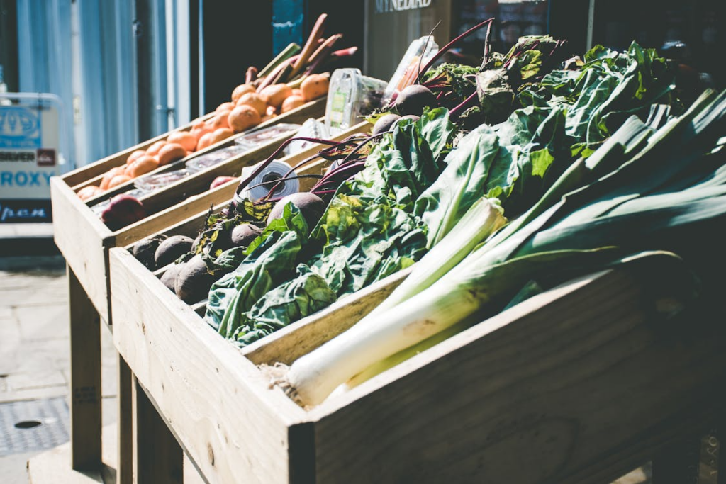 Wooden crates of fresh vegetables at an outdoor market, including leeks, beets, onions, and leafy greens.