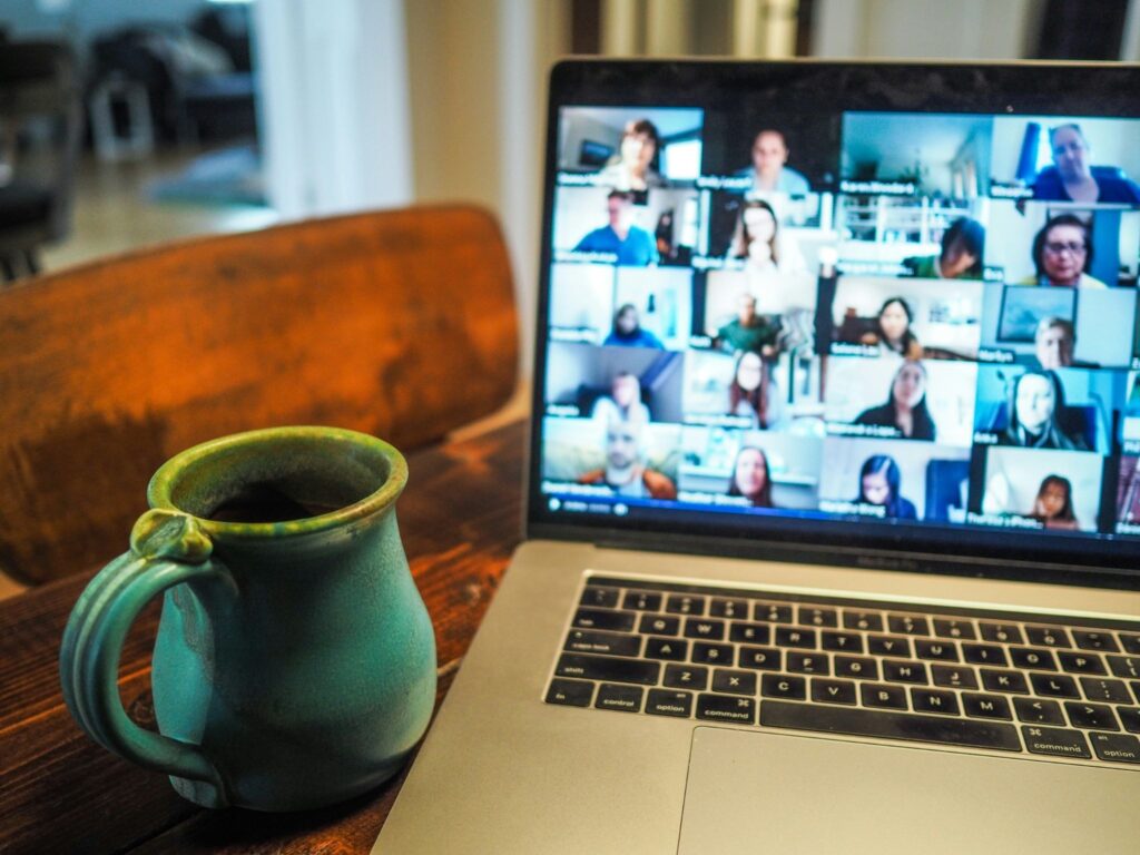 Teal ceramic mug on a wooden table in the foreground with a laptop showing a grid of people in a video conference in the background