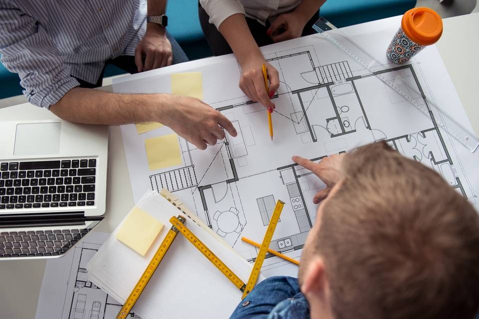 Three professionals in an office point to various sections of a large blueprint on a table beside an open laptop. - Lead Grow Develop - Business, Technology, Personal Development Three professionals in an office point to various sections of a large blueprint on a table beside an open laptop.