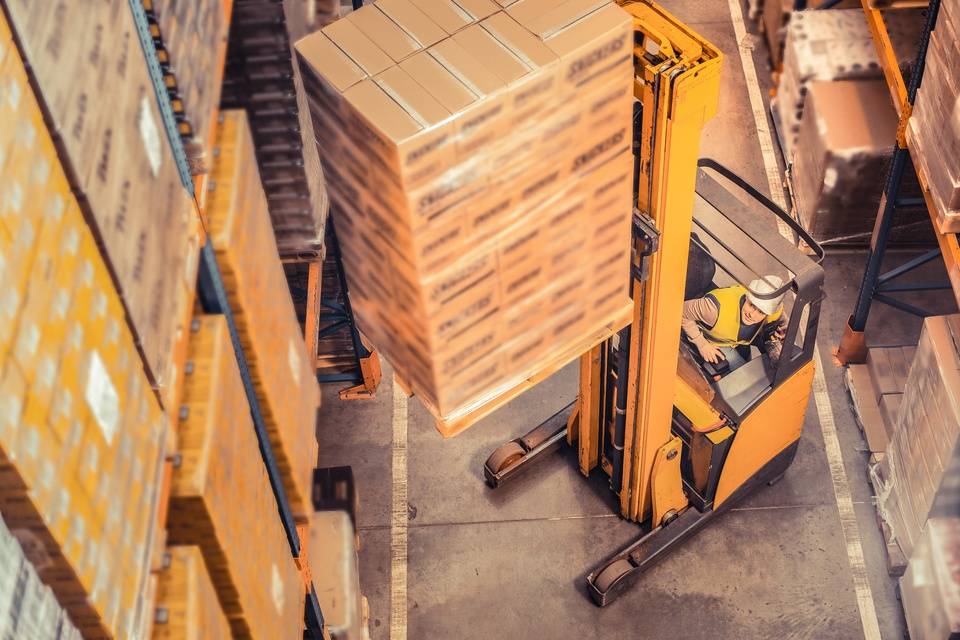 A warehouse worker operates a forklift to lift a large pallet of boxed inventory between tall storage racks.