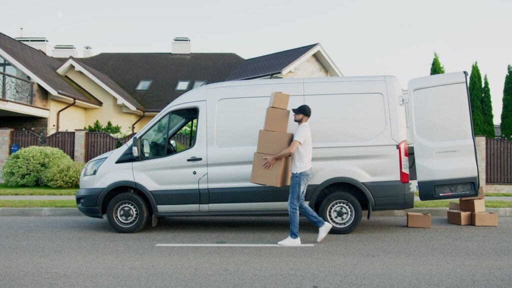 image - Lead Grow Develop - Business, Technology, Personal Development A mover carries a tall stack of cardboard boxes toward the open rear doors of a white transit van parked on a residential street