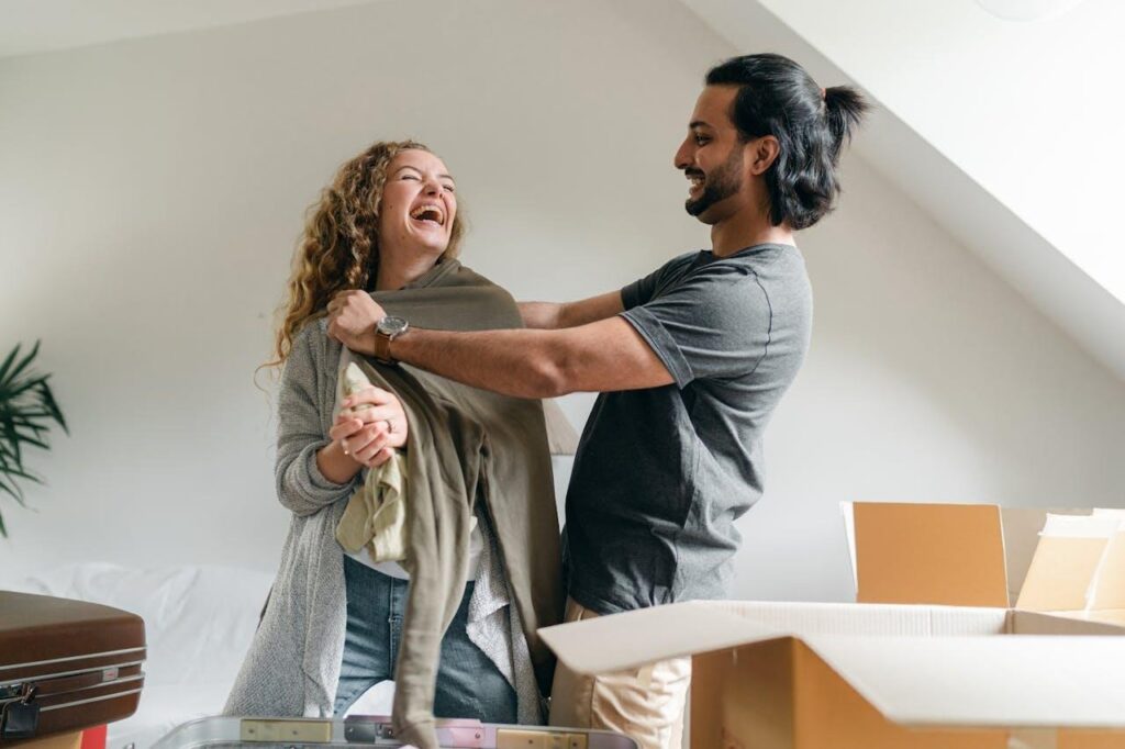 image - Lead Grow Develop - Business, Technology, Personal Development A laughing couple stands together among open moving boxes in their new home, one holding up a piece of clothing they unpacked
