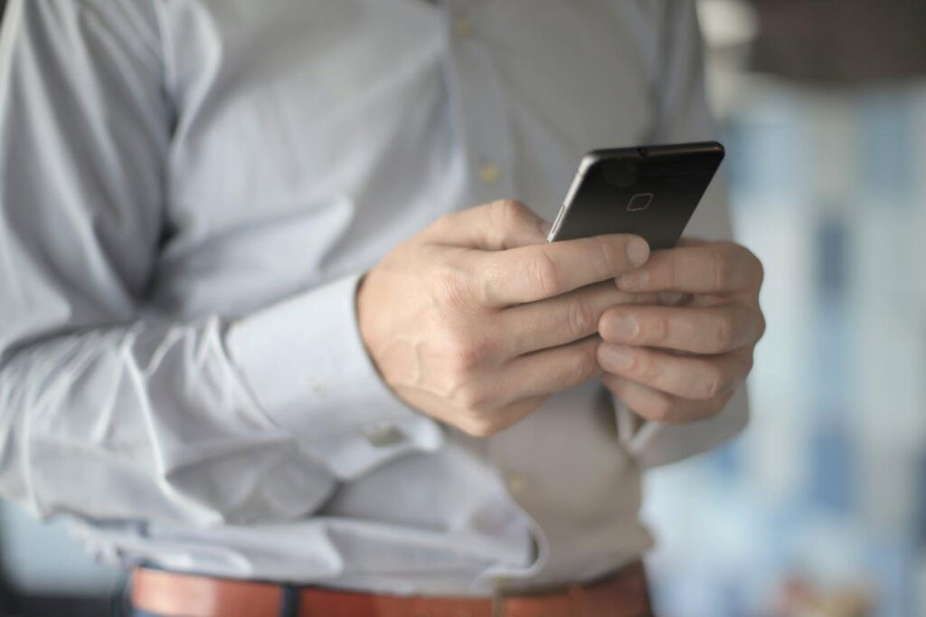 image - Lead Grow Develop - Business, Technology, Personal Development Person in a light shirt using a smartphone, held with both hands at chest level in a bright indoor setting.
