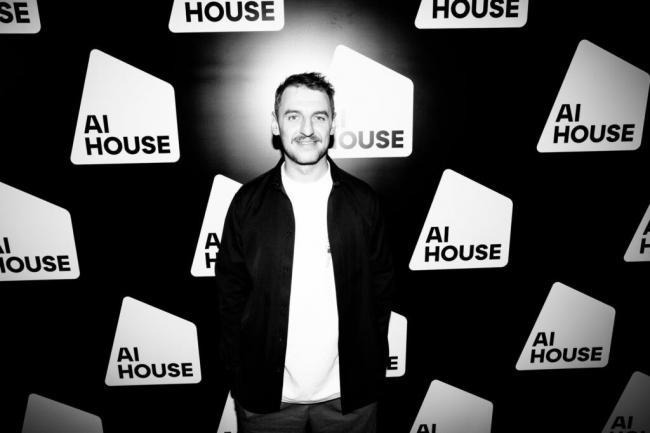 image - Lead Grow Develop - Business, Technology, Personal Development Black-and-white photo of a man posing in front of an AI House step-and-repeat backdrop with repeated logos on a black background and glow behind him.