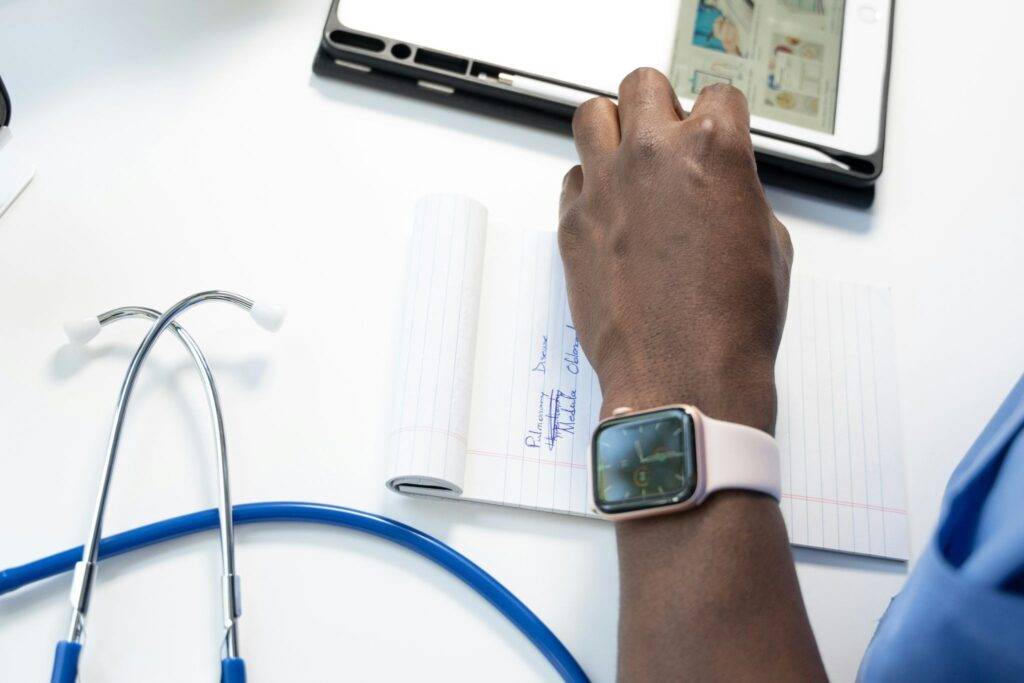 image - Lead Grow Develop - Business, Technology, Personal Development Close-up of a hand wearing a pink smartwatch, with a stethoscope on a white desk and a notebook and tablet nearby on the surface.