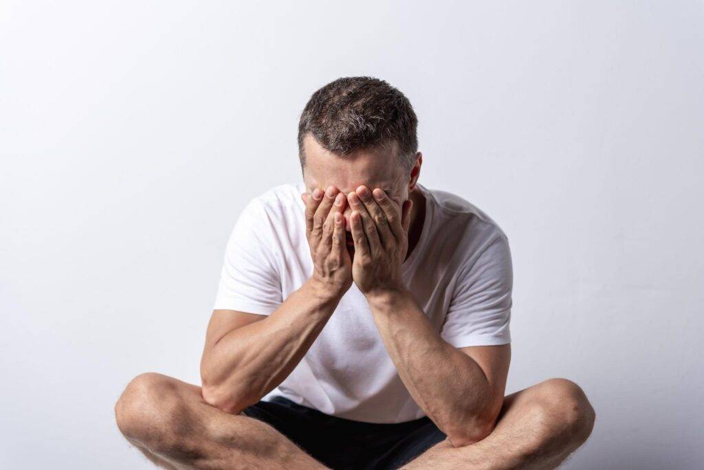 image - Lead Grow Develop - Business, Technology, Personal Development Man sitting cross‑legged in a white shirt, hands covering his face in apparent distress or sadness amid a plain background.
