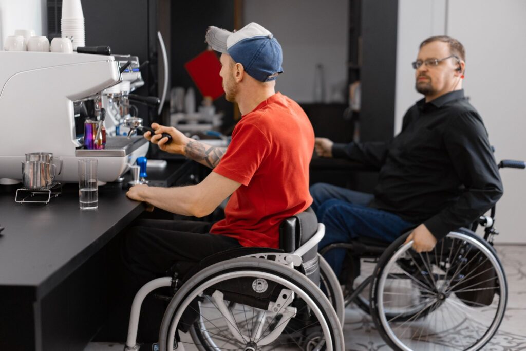 Two men in wheelchairs at a cafe counter; one wearing a red shirt operates an espresso machine, the other watches from nearby.