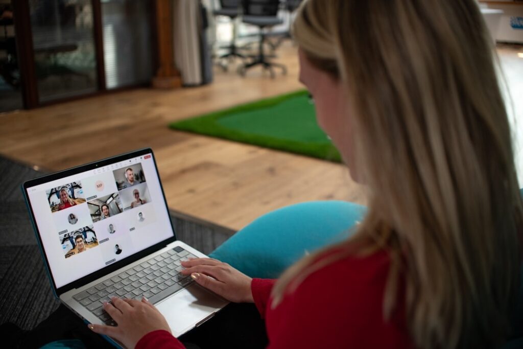 Person with blonde hair in a red sweater using a laptop for a video conference, tiles of participants visible on the screen.