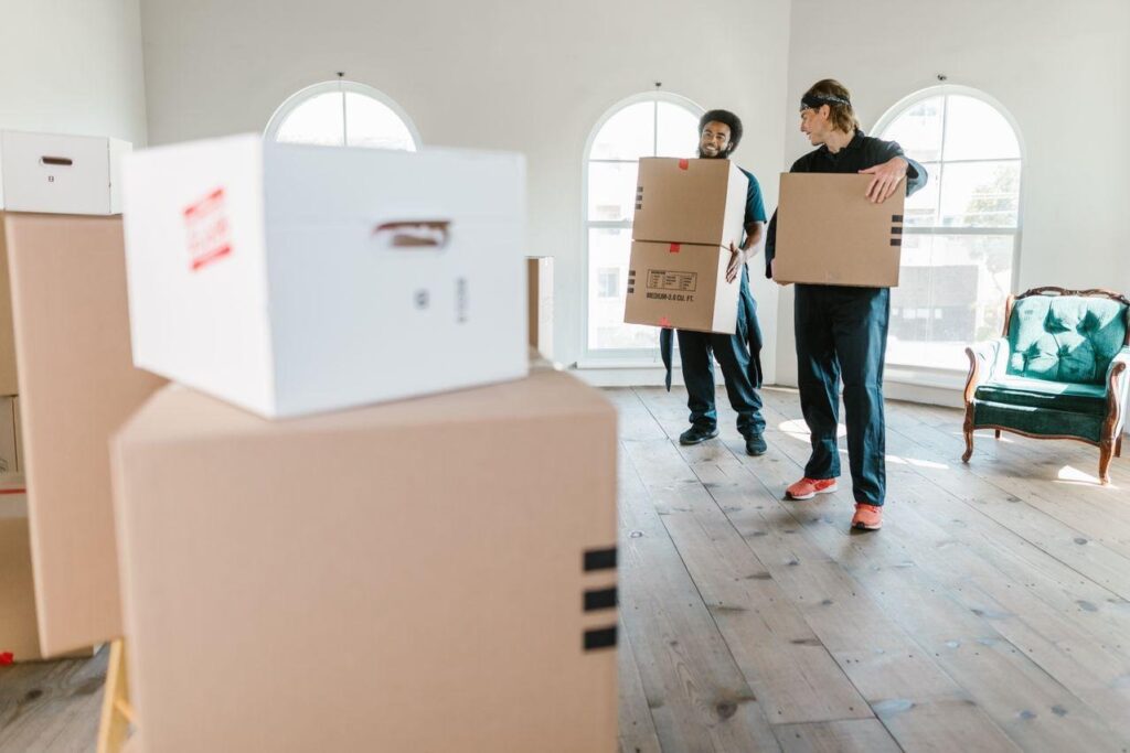 image - Lead Grow Develop - Business, Technology, Personal Development Two movers carry large cardboard boxes in a sunlit room with arched windows and a green chair to the side.