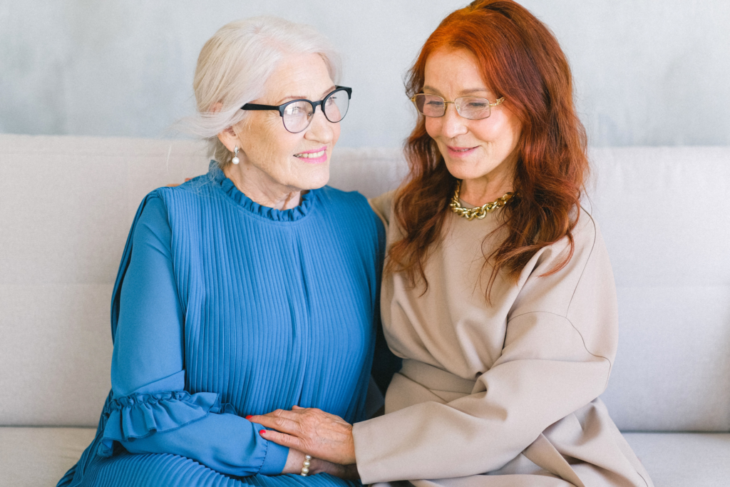 image - Lead Grow Develop - Business, Technology, Personal Development Two women sit close together on a sofa, smiling; older woman in a blue dress with glasses, younger woman in beige with glasses and a gold necklace.