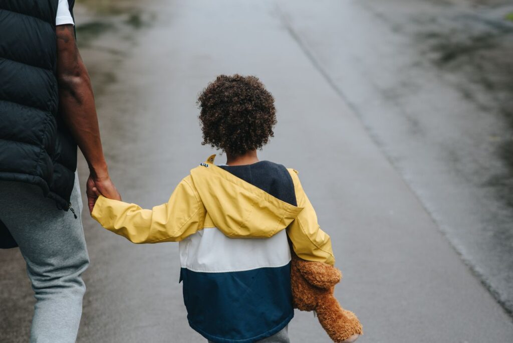Father and child walking together outdoors, holding hands, with a teddy bear in hand. Back view on a wet road