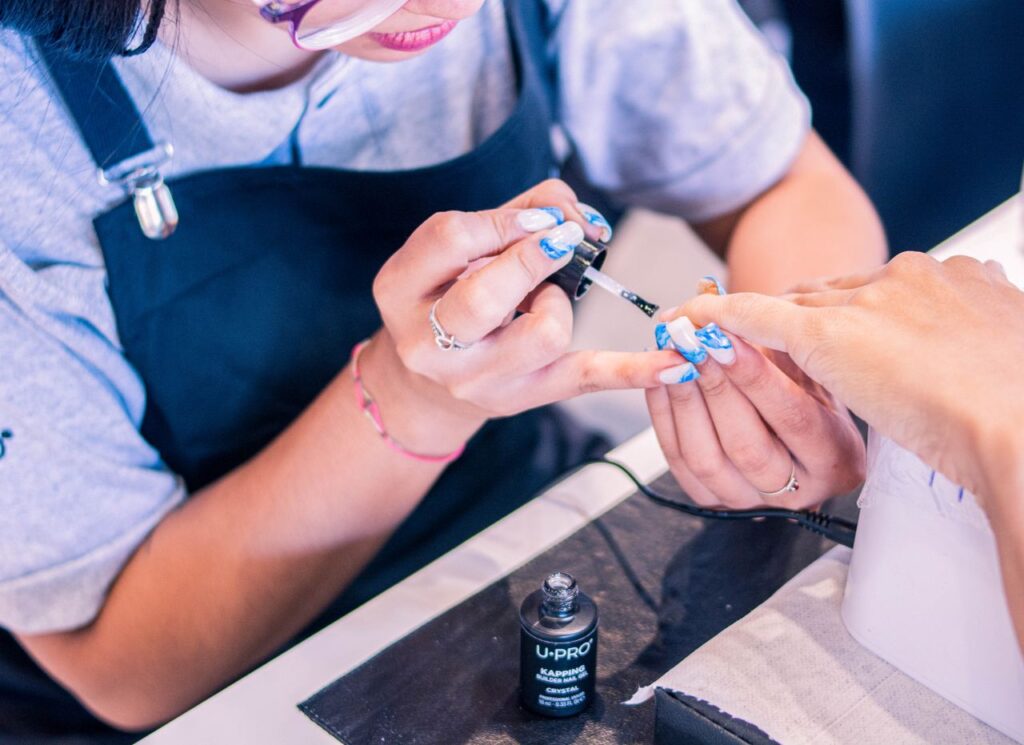 Manicurist applies blue nail polish to client's nails using a brush, with the polish bottle on the table nearby.
