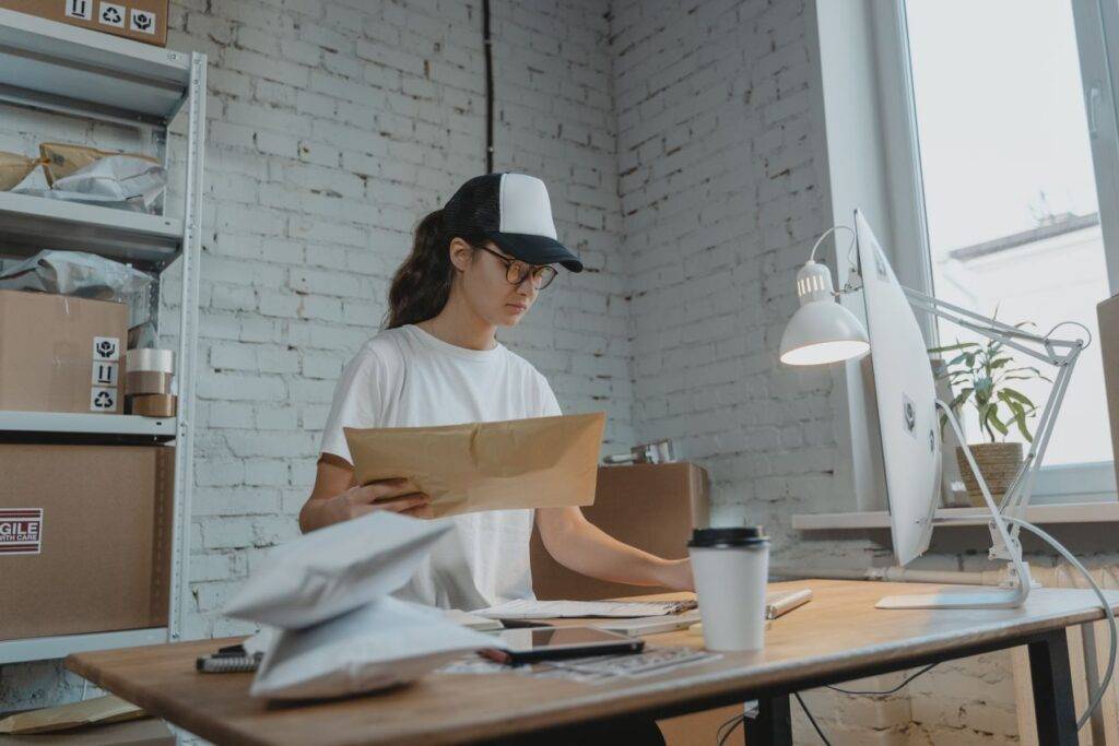 Woman in a white t‑shirt and cap reads a large brown envelope at a cluttered desk in a bright studio/office with a computer and coffee cup nearby