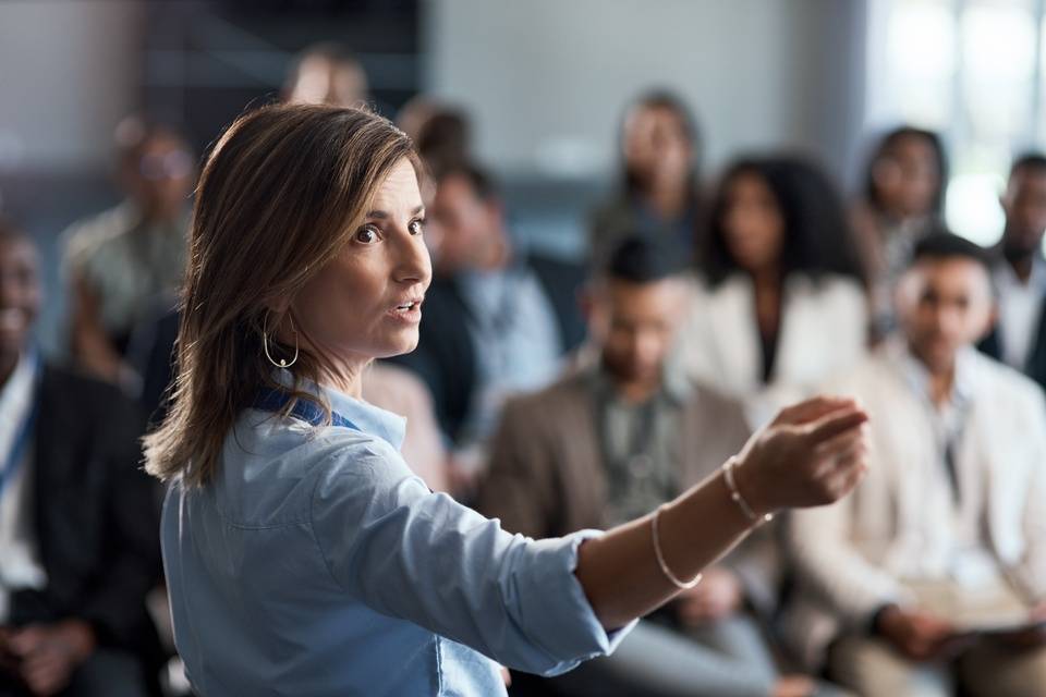 A woman in a blue shirt pointing towards something while speaking to a large group of business professionals.
