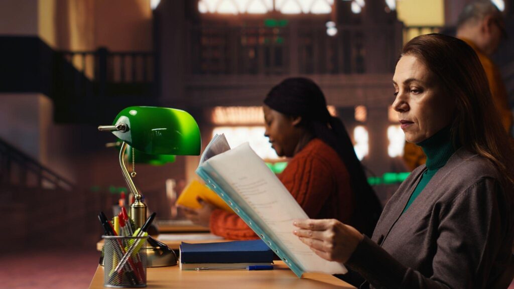 image - Lead Grow Develop - Business, Technology, Personal Development Woman in a blazer reads a stack of documents at a desk, with a green desk lamp and office supplies nearby.