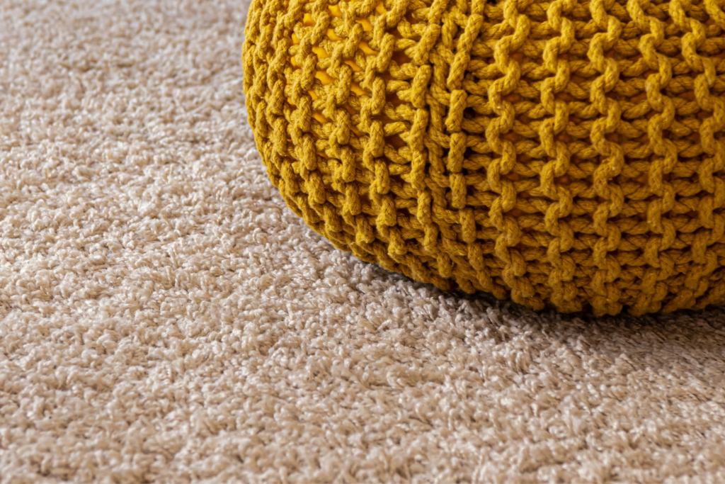 Mustard-yellow chunky knitted pouf resting on a beige carpet.