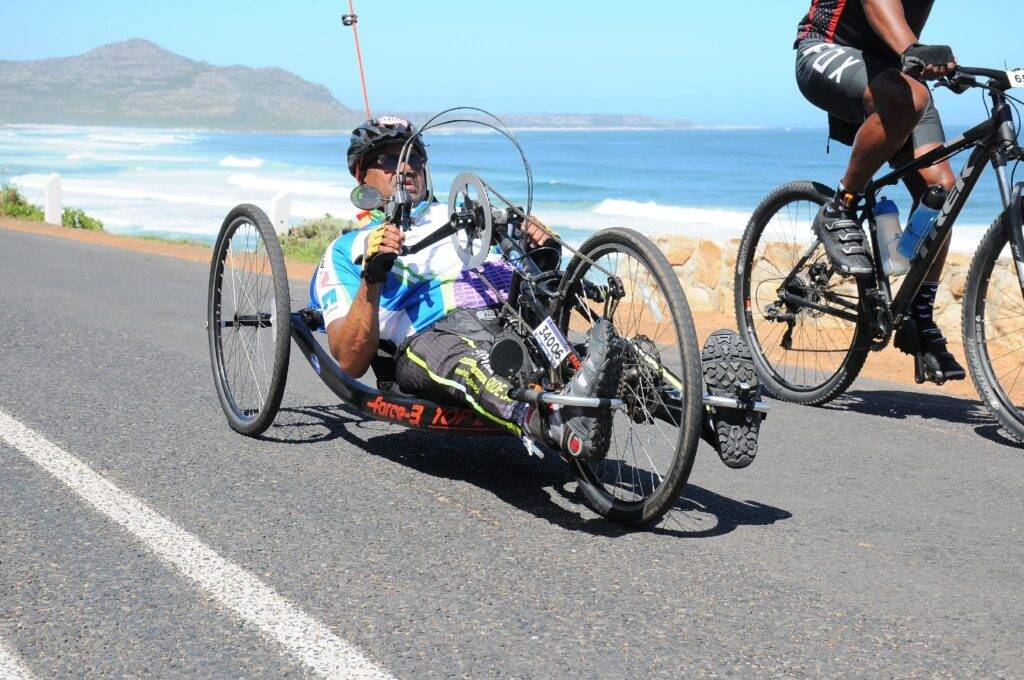 Man in a handcycle racing along a coastal road, cyclist in the background with ocean and mountains visible behind.