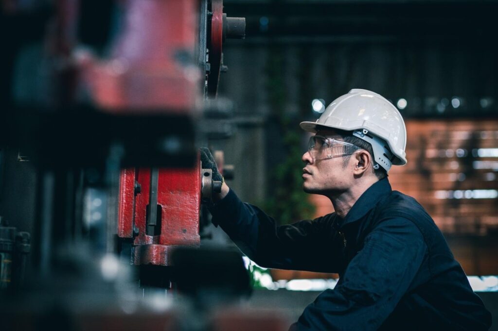 Man wearing a hard hat and safety glasses operating a large red machine in an industrial setting, focusing intently on the controls.