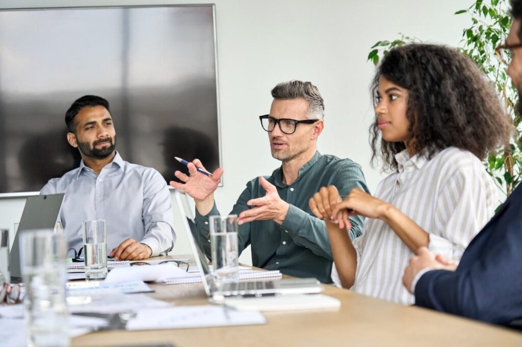 Four professionals in a modern meeting room discuss a project around a table with laptops and documents on display.
