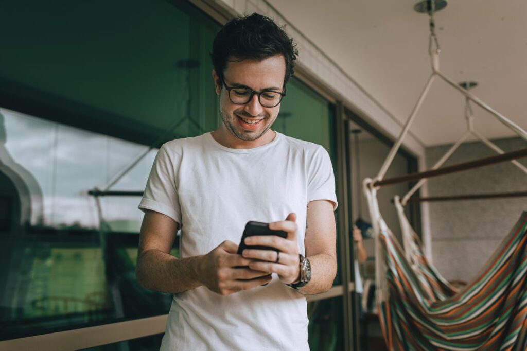 Man in a white tee and glasses smiles at his phone on a sunny balcony with a striped hammock in the background.
