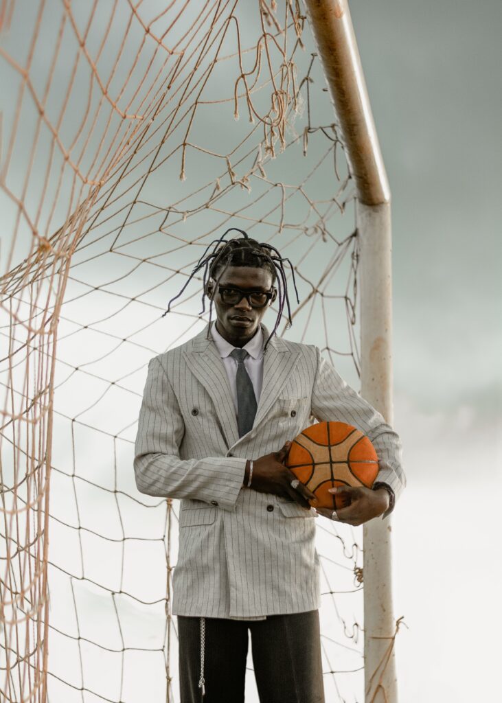 Man in a gray pinstripe suit holding a basketball, standing in front of a torn goal net outside.