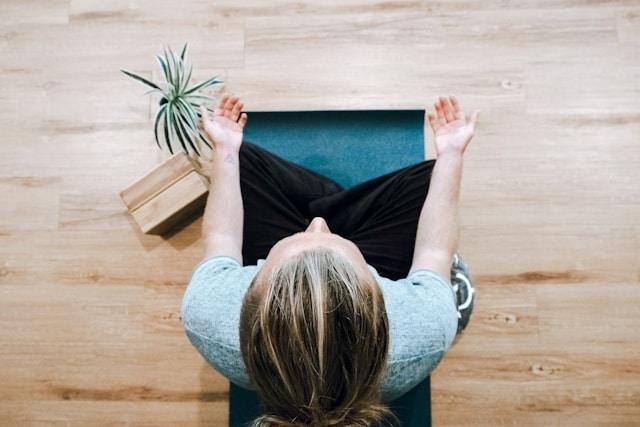 image - Lead Grow Develop - Business, Technology, Personal Development Overhead view of a person seated on a teal yoga mat, arms extended outward, preparing for a stretch, with a small plant and wooden floor nearby.