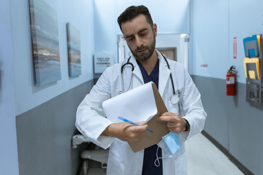 Male doctor in a white coat in a hospital corridor, examining a clipboard with a stethoscope around his neck.