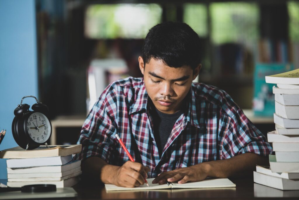 image - Lead Grow Develop - Business, Technology, Personal Development Young man in a plaid shirt writes in a notebook at a desk surrounded by stacks of books and a vintage alarm clock nearby.