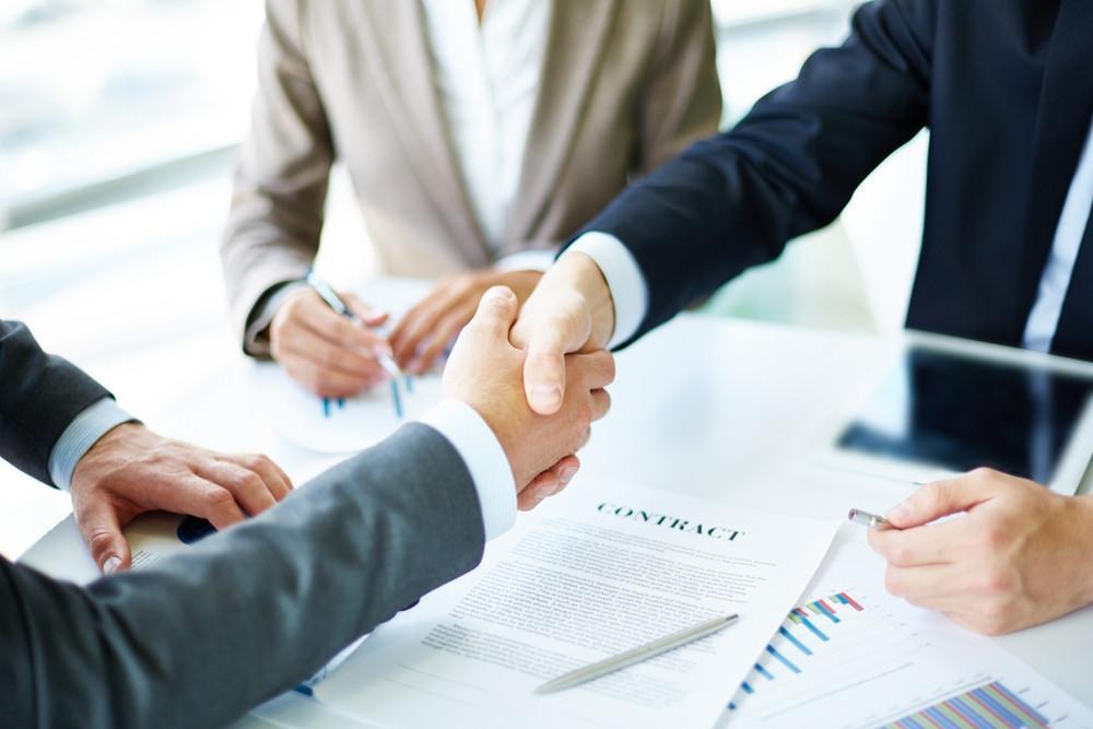 image - Lead Grow Develop - Business, Technology, Personal Development Two professionals shake hands over a signed contract on a desk, signaling an agreement.