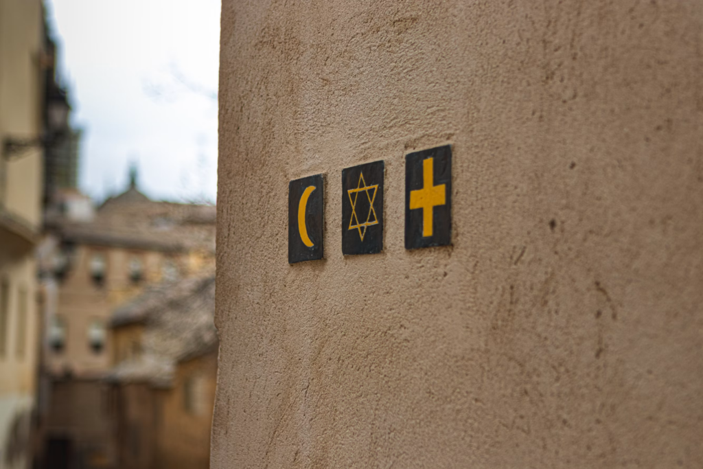 Three dark tiles mounted on a beige wall show a yellow crescent, a Star of David, and a cross in an alley.