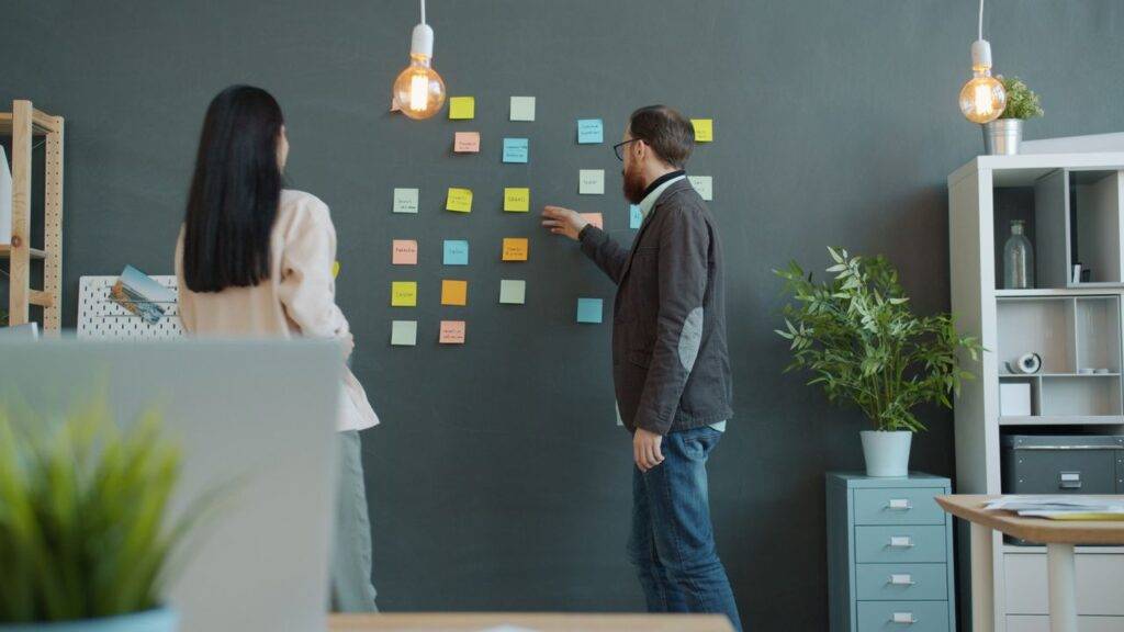 Two adults collaborate on a project using sticky notes on a blackboard.