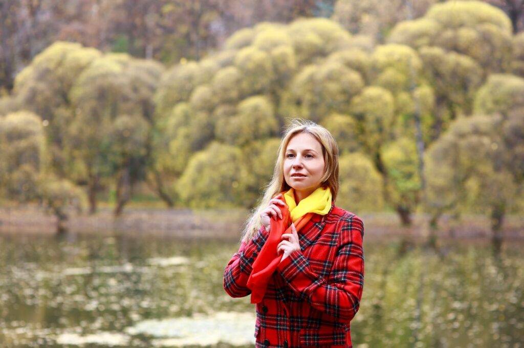 Woman in plaid coat by a serene lake during autumn, showcasing vibrant fashion.